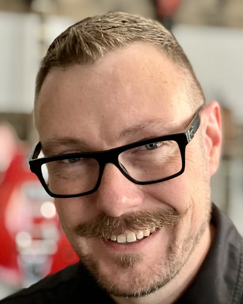 Joshua C. Carroll, smiling, in glasses, with guitars hanging on the wall behind him